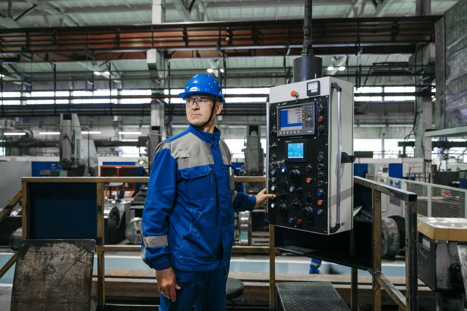 A man wearing a blue overcoat and hard hat stands in a factory pressing a button on an industrial machine
