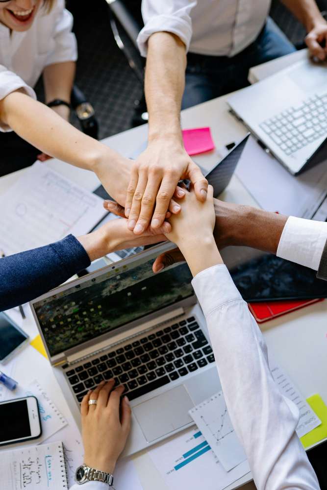 5 arms outstretched to meet in the middle of different races and genders above laptops on a desk