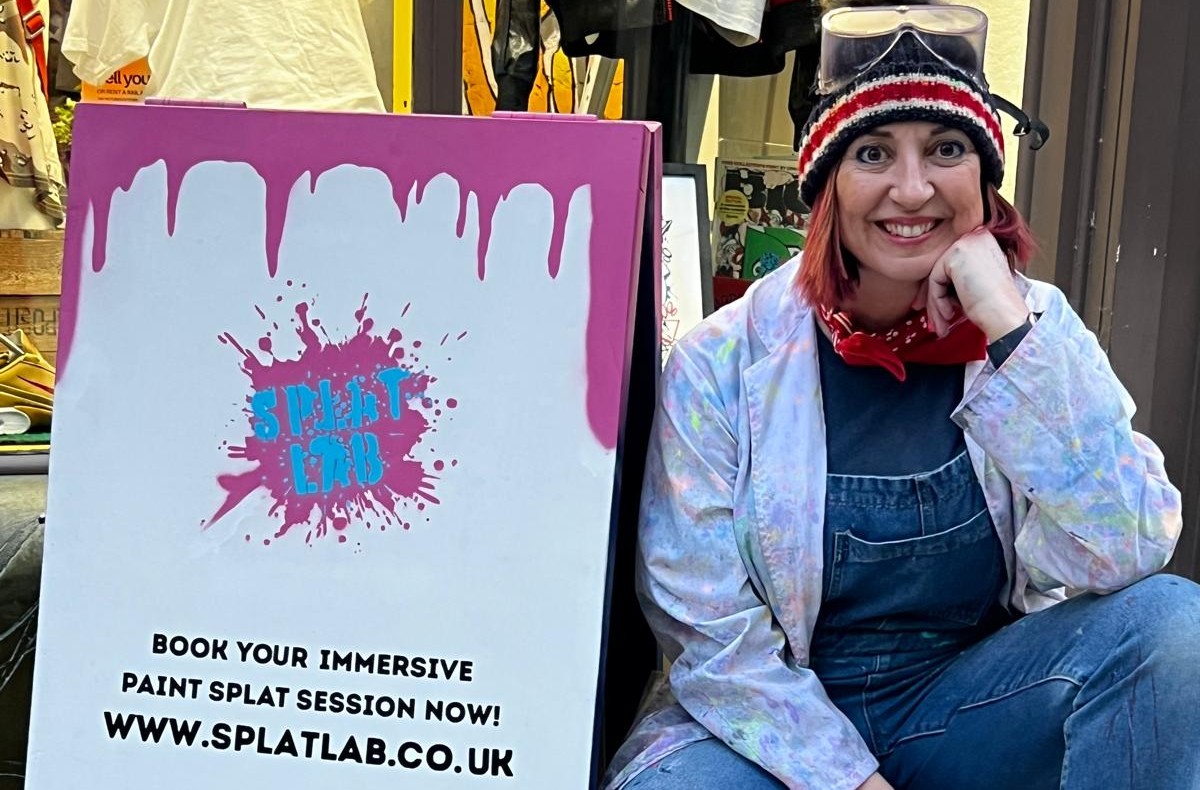 Sonja Morgan, owner of SplatLab kneeling down next to a SplatLab sandwich sign
