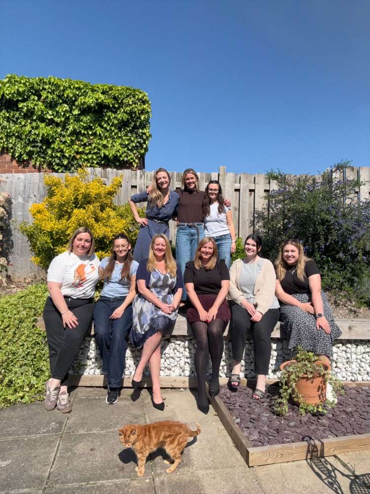9 women sit and stand in a garden having their photo taken, a ginger cat stands at the front